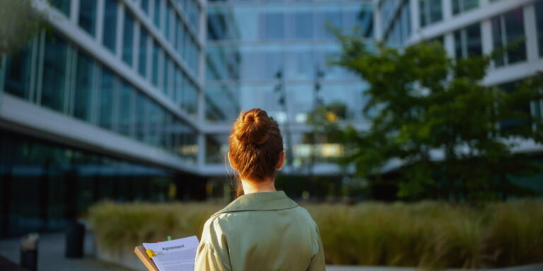 A woman in a light-colored coat stands outside a modern glass office building, holding a clipboard with an agreement document. The greenery and well-maintained landscape suggest a focus on property upkeep and management. This image represents the importance of facilities maintenance in ensuring clean, functional, and professional-looking commercial properties.